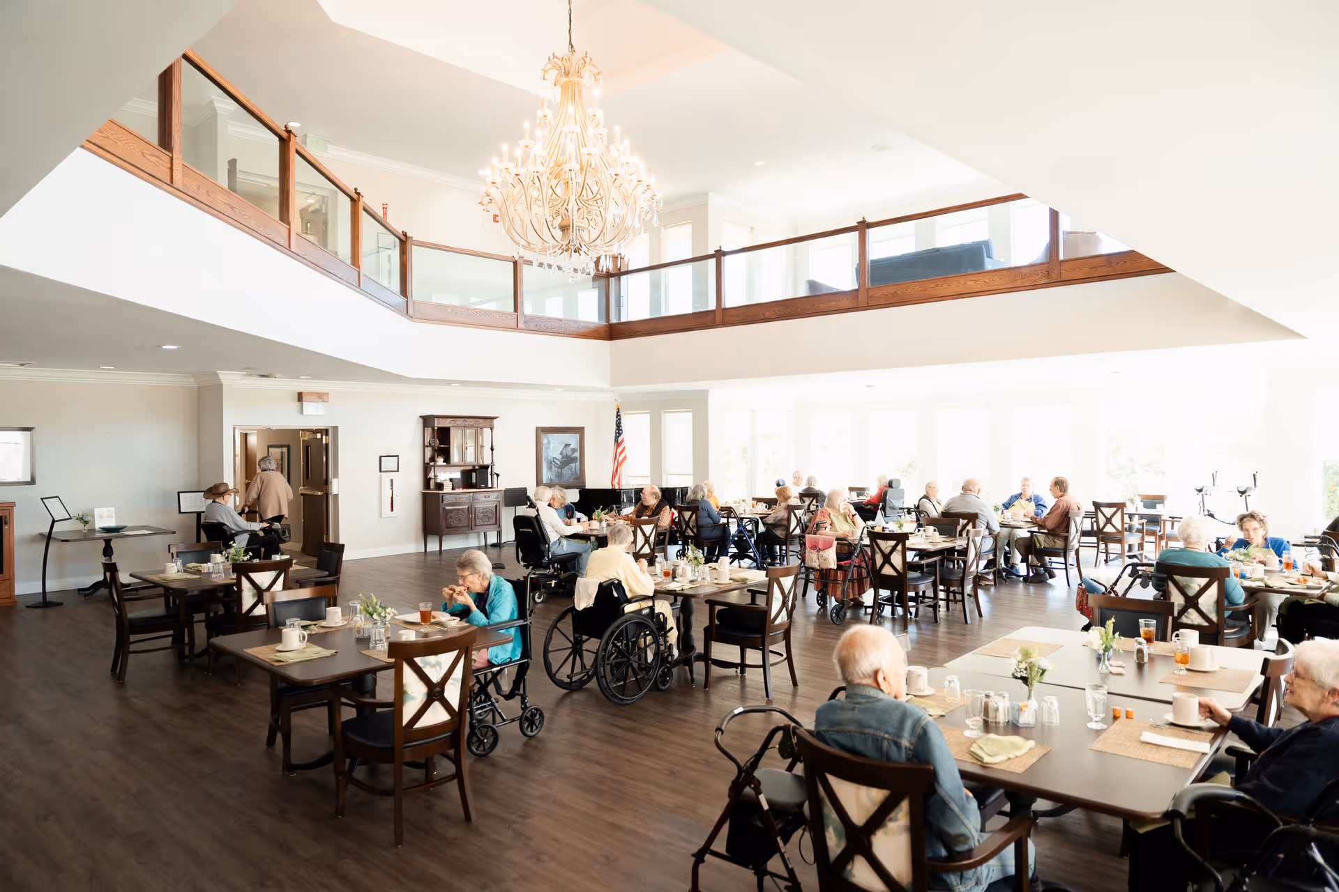 Bright, open dining room in a senior living facility with residents seated at tables, a large chandelier, and an upstairs mezzanine.