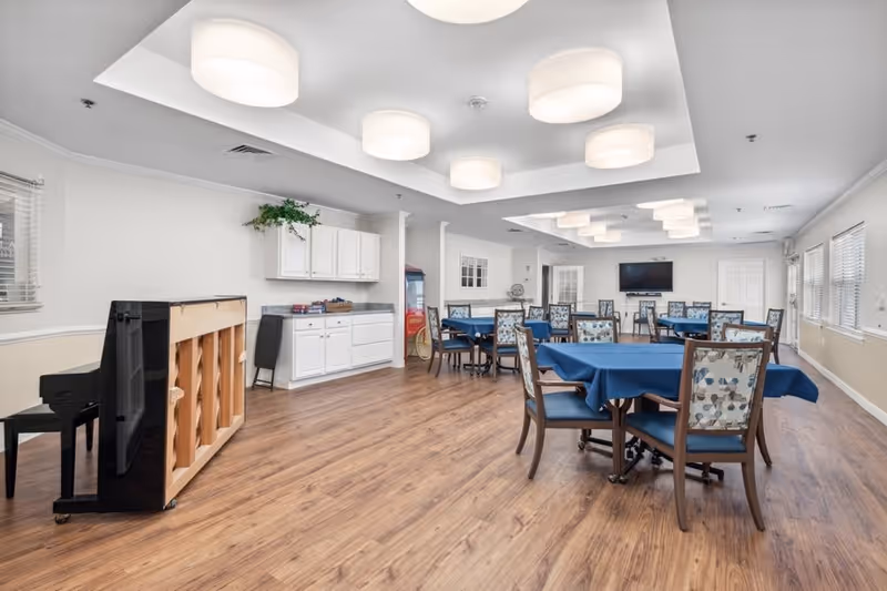 A spacious dining room with several tables covered in blue tablecloths and chairs with floral patterned backs. The room has wood flooring, white walls, and large windows on one side. There is a black upright piano on the left side and white cabinets with a countertop along the back wall. Ceiling lights are modern and circular, evenly spaced across the ceiling. A flat-screen TV is mounted on the far wall.