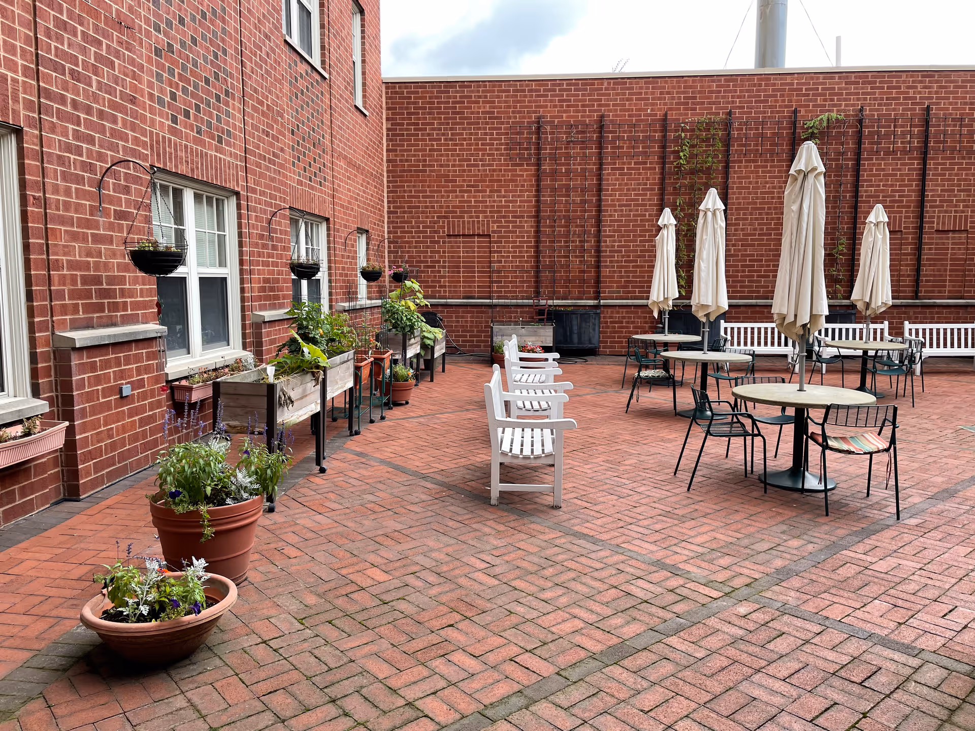 Outdoor patio area with brick flooring and brick walls, featuring white benches, round tables with closed beige umbrellas, black metal chairs, and various potted plants and hanging planters along the wall.