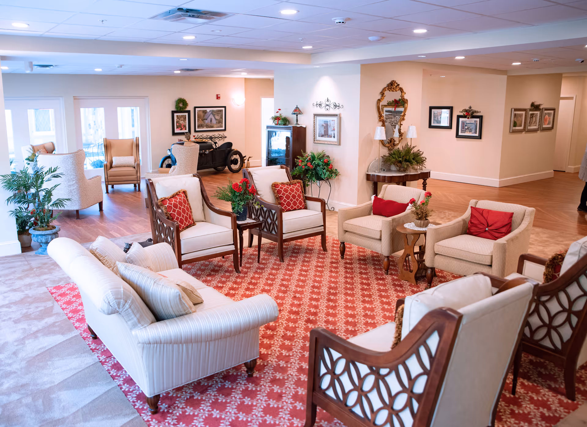 A spacious and well-lit senior living facility common area with comfortable seating including sofas and armchairs arranged around a red patterned rug. The room features wooden flooring, decorative plants, framed artwork on the walls, and a vintage car displayed in the background. There is also a decorative mirror and table with lamps and plants, creating a warm and inviting atmosphere.