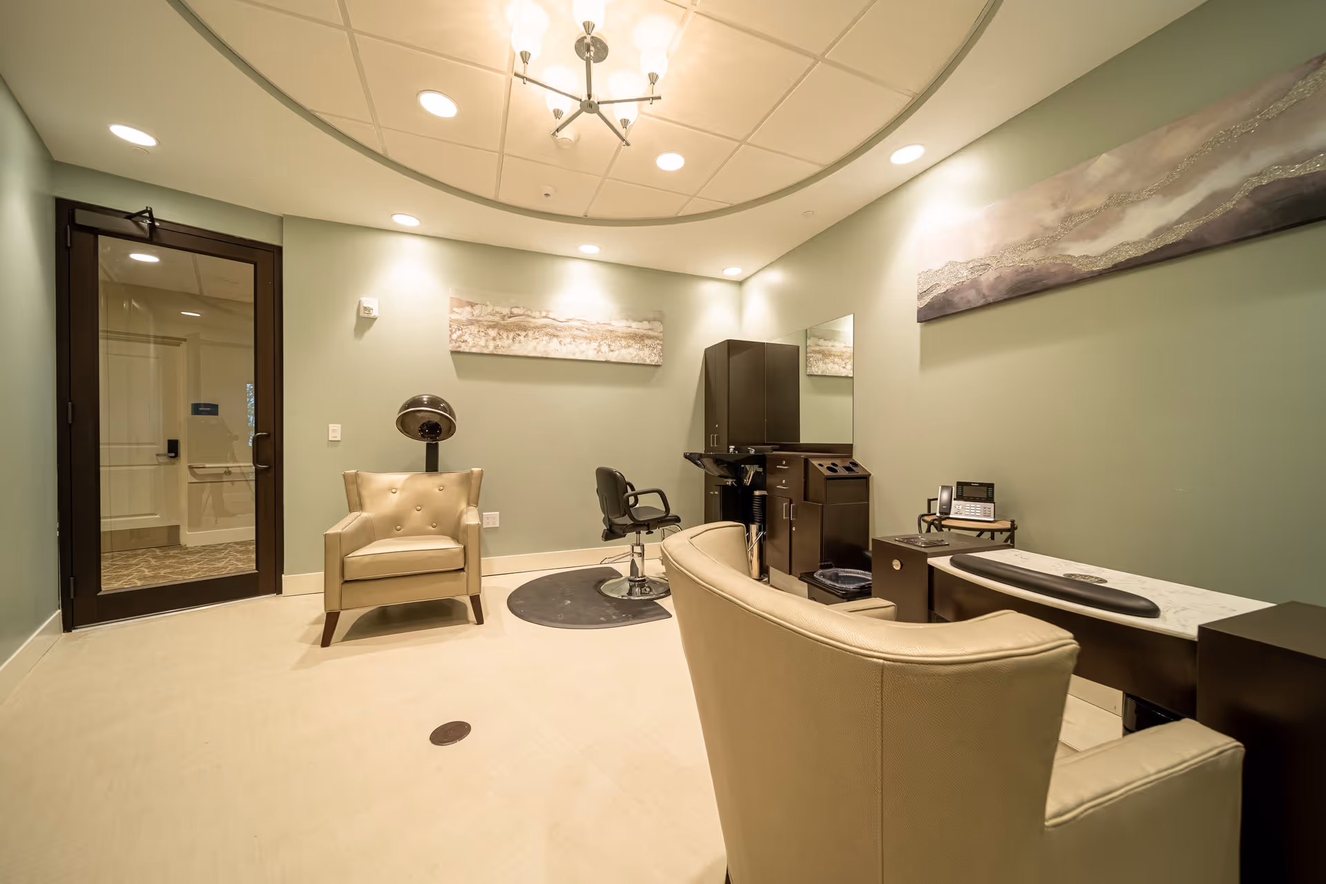 A well-lit salon room with light green walls, featuring a beige armchair, a black salon chair on a circular mat, a hair dryer hood, a dark wood cabinet with a mirror, and a small desk with a phone and other items. The ceiling has recessed lighting and a modern chandelier, and there are two abstract paintings on the walls.