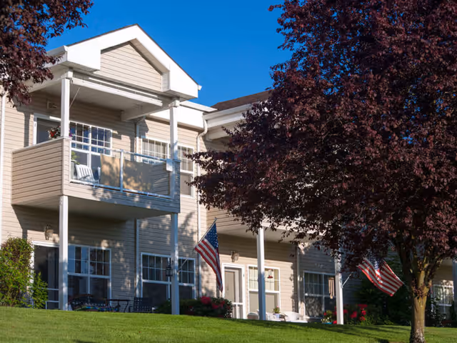 Exterior view of a two-story residential building with beige siding, balconies, and American flags displayed near the entrances. There are trees with dark red leaves and a well-maintained green lawn in front of the building under a clear blue sky.