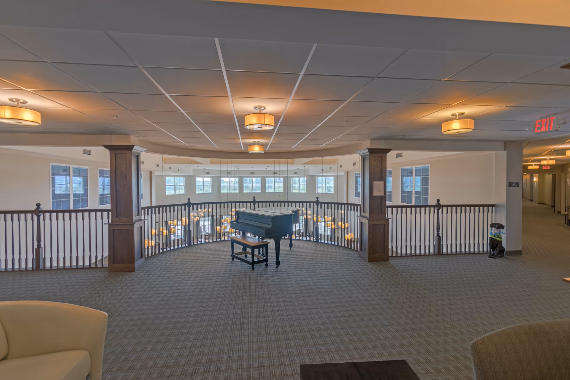 Interior view of a senior living facility showing a spacious carpeted area with a black grand piano and bench in the center. The area is surrounded by a wooden railing overlooking a lower floor with hanging lights. There are large windows letting in natural light, ceiling lights, and a hallway with an exit sign. A small decorative dog statue wearing a green scarf is near one of the pillars.