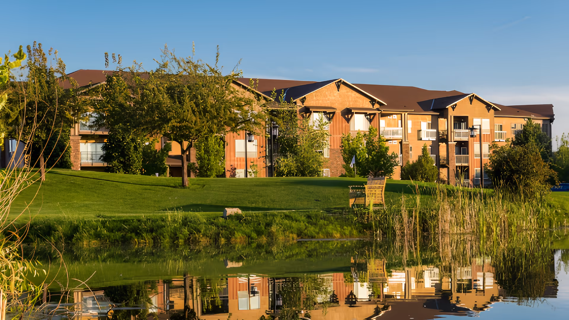 A senior living facility building with a brown roof and multiple windows, surrounded by green trees and a well-maintained lawn. In the foreground, there is a pond reflecting the building and greenery, with a wooden bench near the water's edge under a clear blue sky.