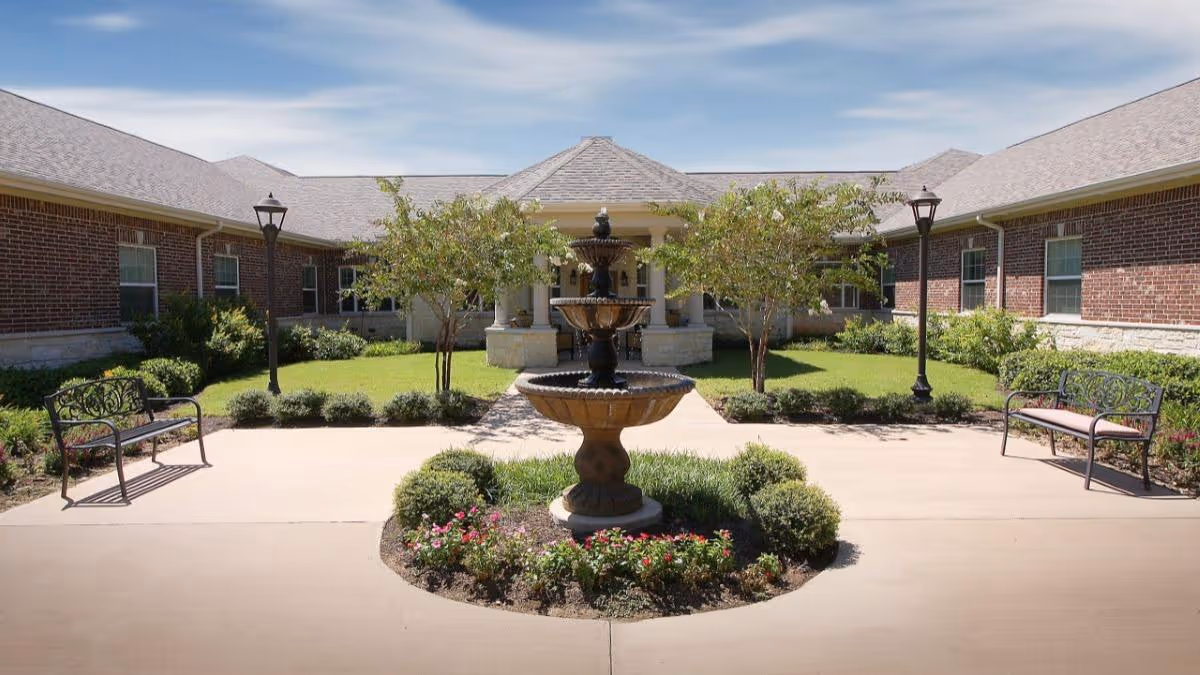 Outdoor courtyard area at Anointed Senior Living Austin featuring a three-tiered water fountain surrounded by a circular flower bed with shrubs and flowers. There are two benches on either side of the paved walkway, lamp posts, and brick buildings with windows in the background under a partly cloudy sky.