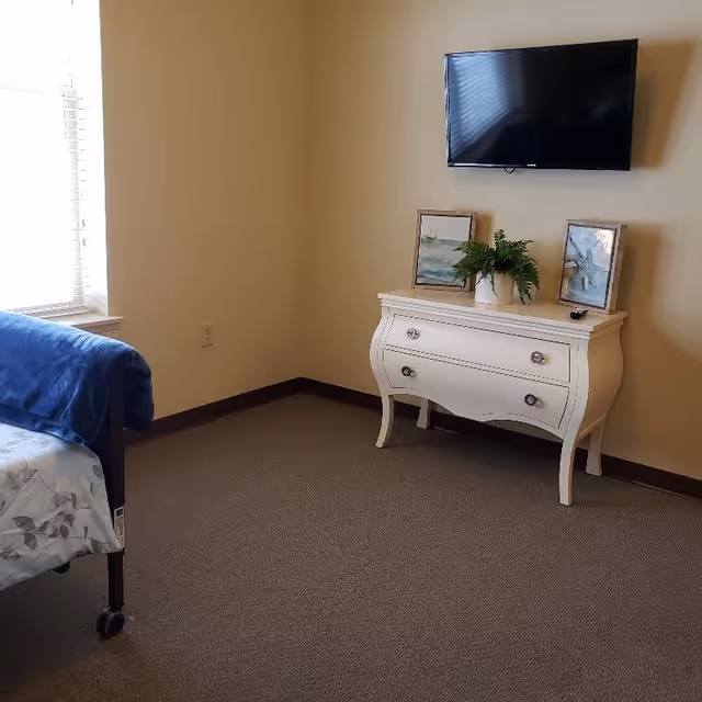 A corner of a senior living bedroom with a bed partially visible on the left covered with a blue blanket. A white dresser with two drawers stands against a beige wall, decorated with two framed pictures and a potted plant. Above the dresser, a flat-screen TV is mounted on the wall. A window with blinds is on the left side, letting in natural light.