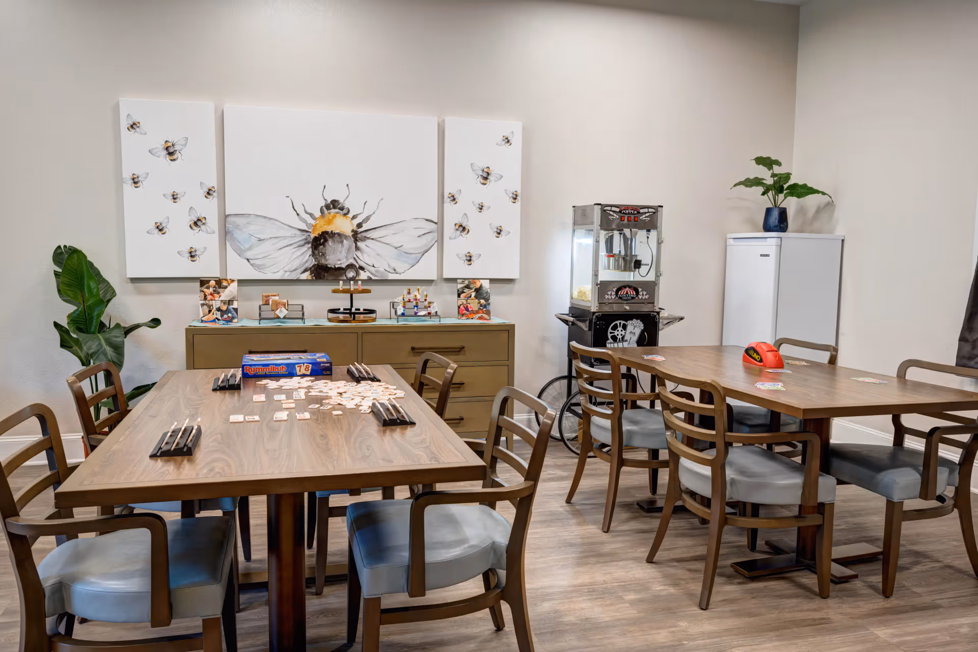 A well-lit activity room with two wooden tables surrounded by chairs. One table has a Rummikub game set up with tiles and racks, while the other table has an Uno card game in progress. On the back wall, there are three bee-themed paintings above a cabinet. To the right, there is a popcorn machine and a white freezer with a potted plant on top. The floor is wooden, and the walls are painted light beige.