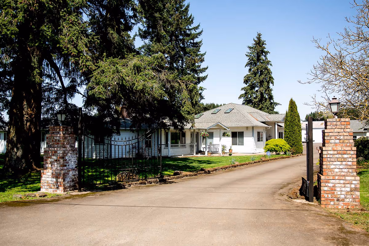 View of a single-story white house with a gray roof, surrounded by tall trees and greenery. The house is accessed by a paved driveway flanked by brick pillars with lanterns on top. The sky is clear and blue.