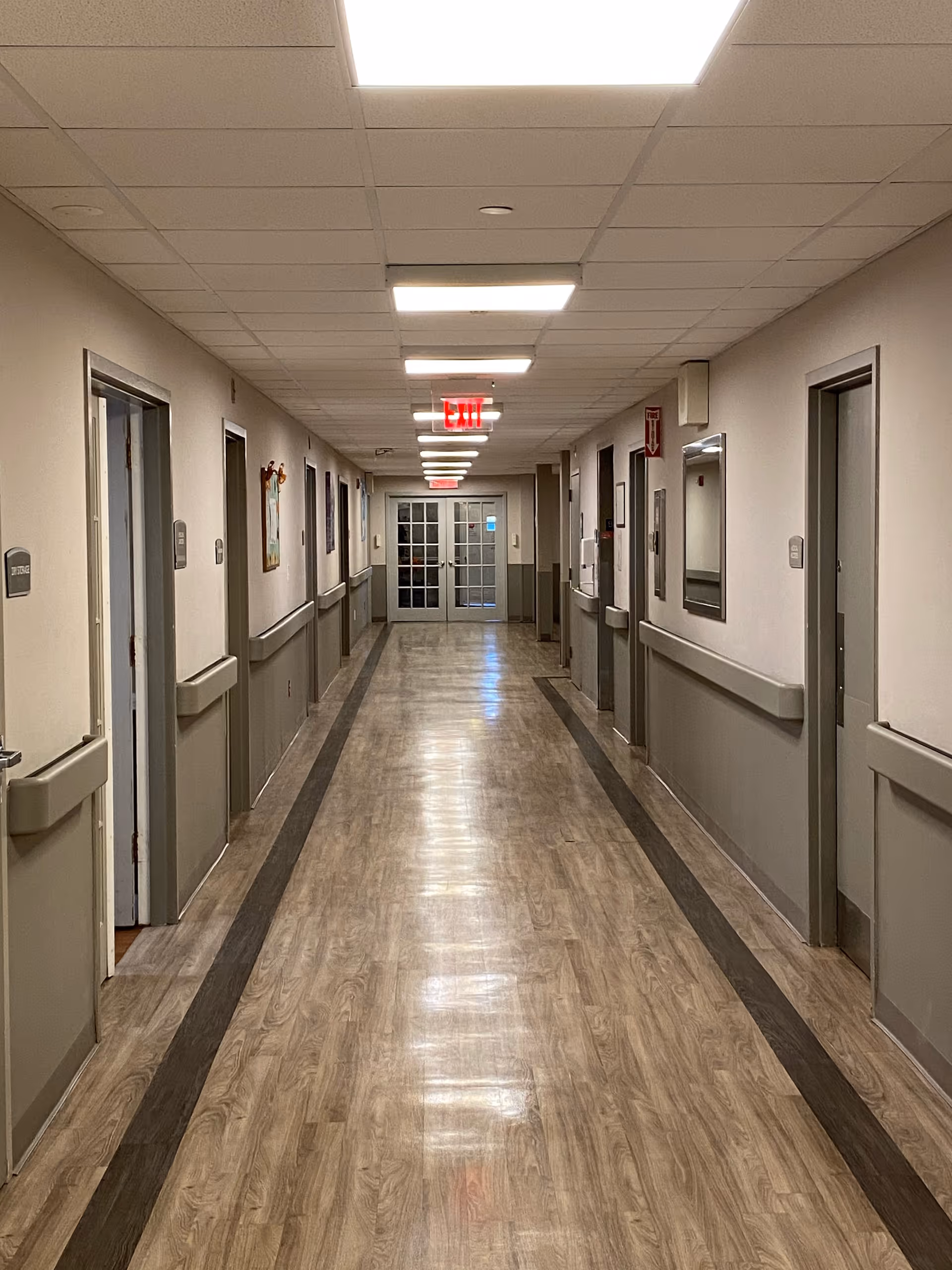 Long, well-lit interior hallway in a senior care facility with doors on both sides, handrails, and double exit doors at the far end.