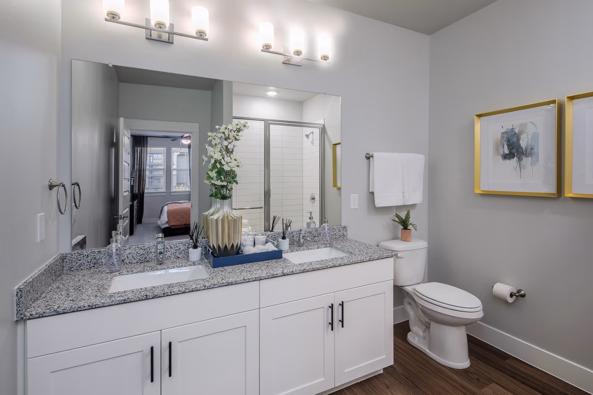 Modern bathroom with a double sink vanity featuring a granite countertop, a large mirror above, and two light fixtures. There is a glass-enclosed shower in the background and a toilet with a small plant on top. Two framed abstract artworks hang on the wall, and a towel rack with white towels is mounted above the toilet. The floor is wood-style.