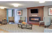 A well-lit communal lounge with armchairs around a coffee table topped with flowers, a wall-mounted TV above a modern fireplace, and windows letting in natural light.