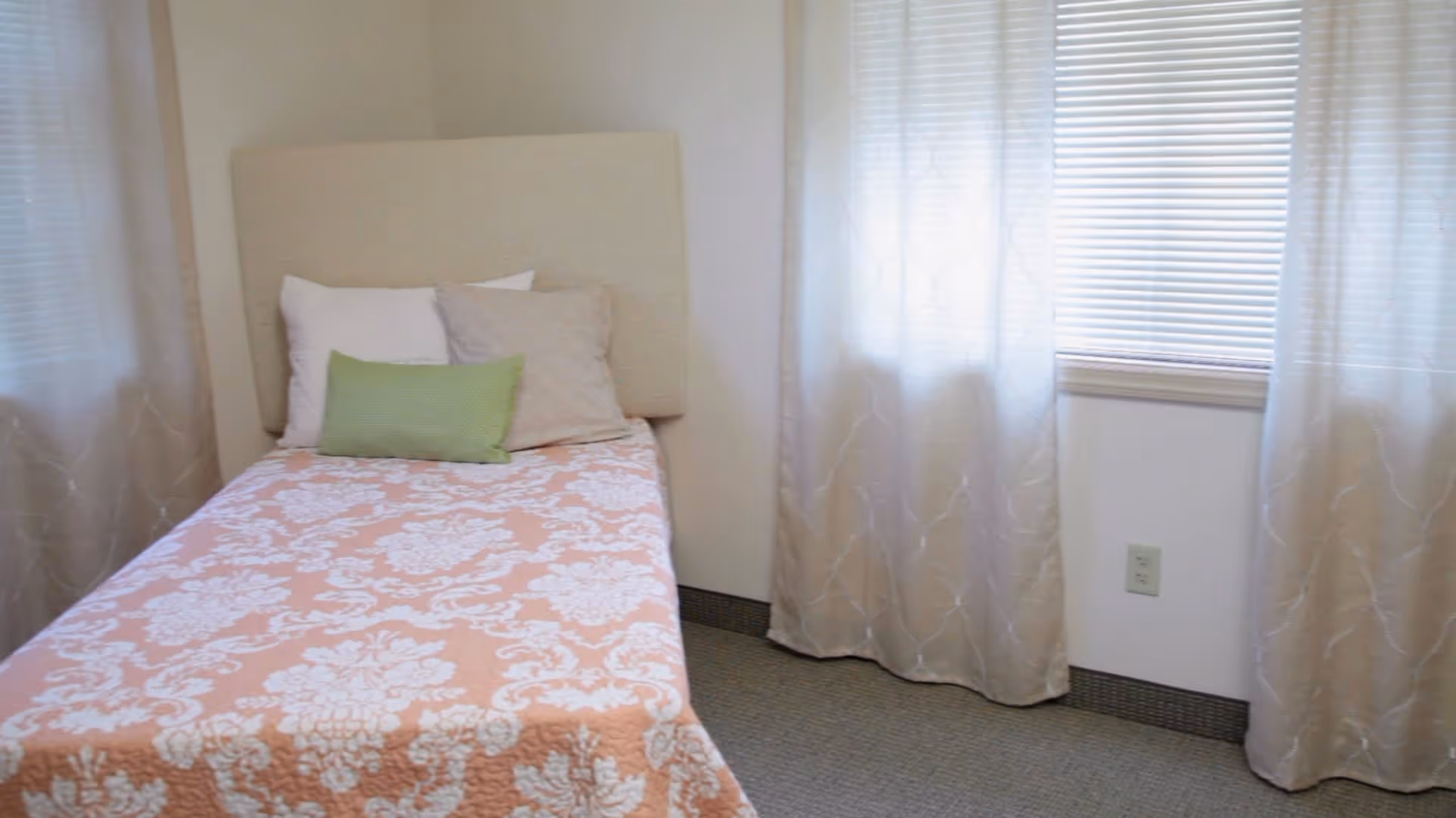 A small bedroom with a single bed covered in a pink and white patterned bedspread. The bed has three pillows in white, beige, and green. The room has light-colored walls, two windows with white blinds and sheer curtains, and a carpeted floor.