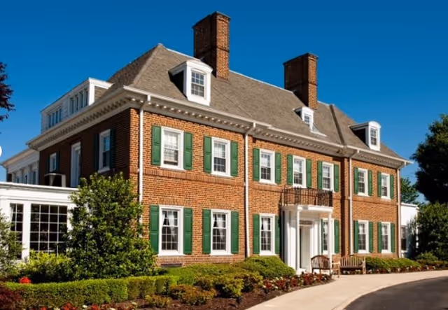 Brick colonial-style building with green shutters and a landscaped front entrance under a clear blue sky.