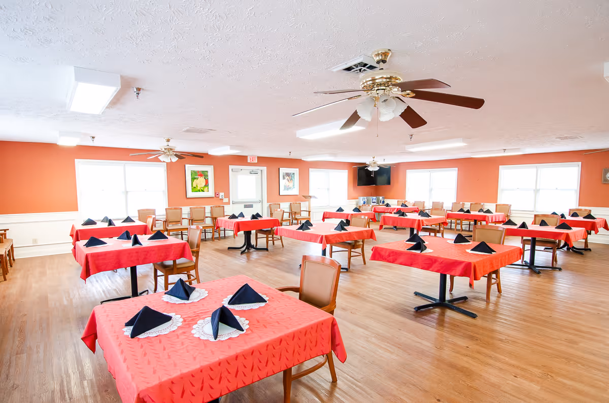 A spacious dining room with multiple tables covered in red tablecloths, each set with black folded napkins on white doilies. The room has wooden flooring, orange walls, several windows letting in natural light, ceiling fans, and chairs arranged around the tables.