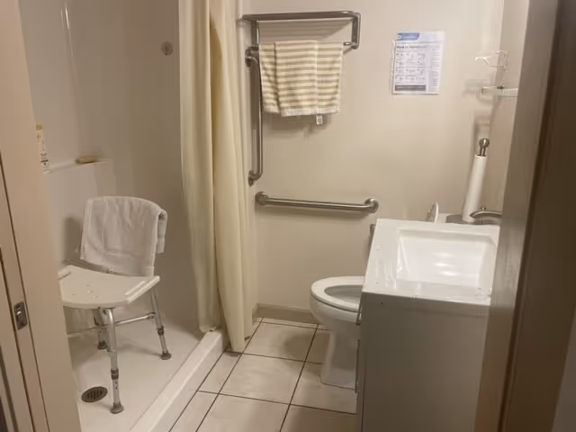 A small bathroom with a walk-in shower equipped with a white shower chair and a white towel hanging on it. There is a beige shower curtain, a toilet, a white vanity with a sink, and a towel rack on the wall holding a striped towel. The floor is tiled and there is a paper towel holder on the vanity.