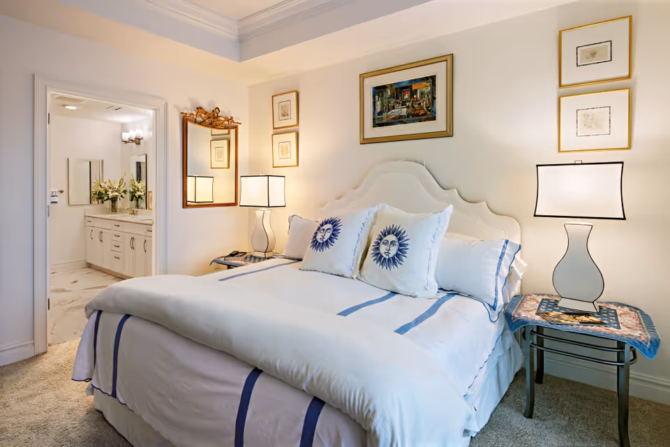 Well-lit bedroom with a neatly made bed featuring sun-themed pillows, bedside tables and lamps, and an open doorway to a bathroom.
