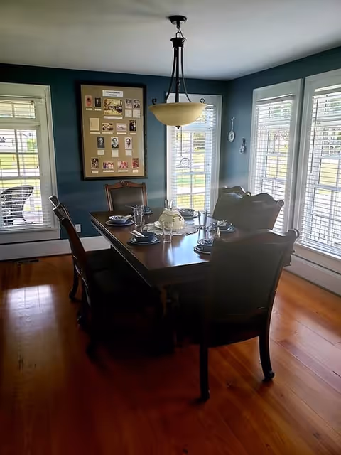 A dining room with a dark wooden table set for six people, surrounded by six matching chairs. The room has teal walls, three large windows with white blinds letting in natural light, and a framed collage of photos and documents on the wall. A hanging light fixture is centered above the table.