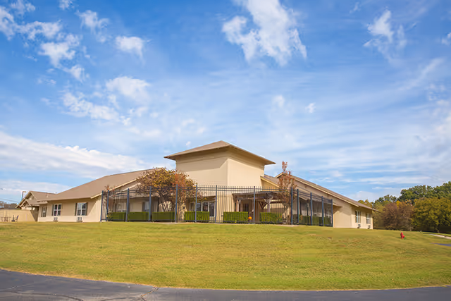 Low single-story beige senior living building with a fenced entrance on a grassy lawn under a blue sky.