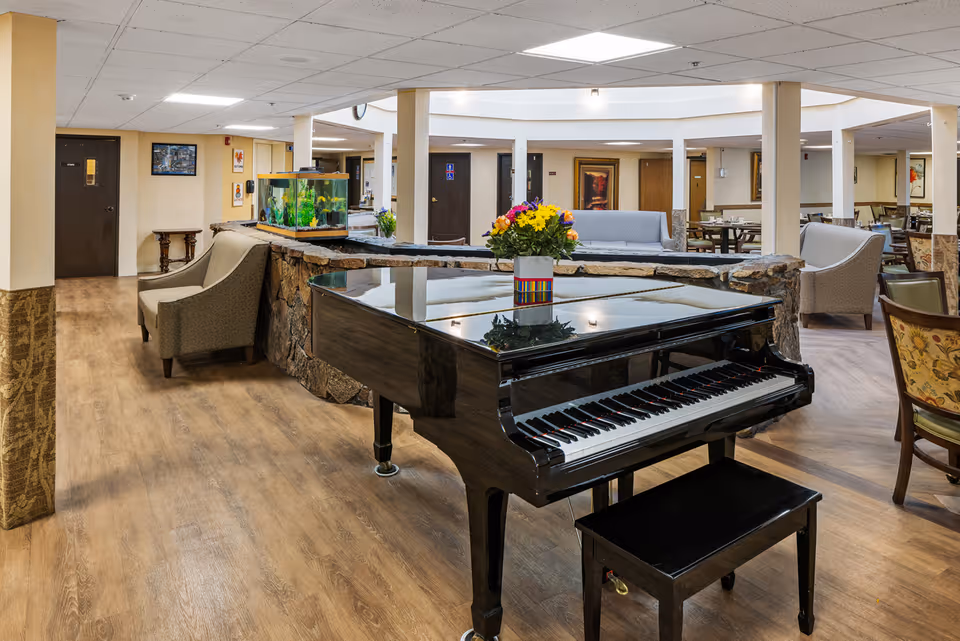 Grand piano with a vase of flowers in a spacious senior living common area with seating, an aquarium, and dining tables.