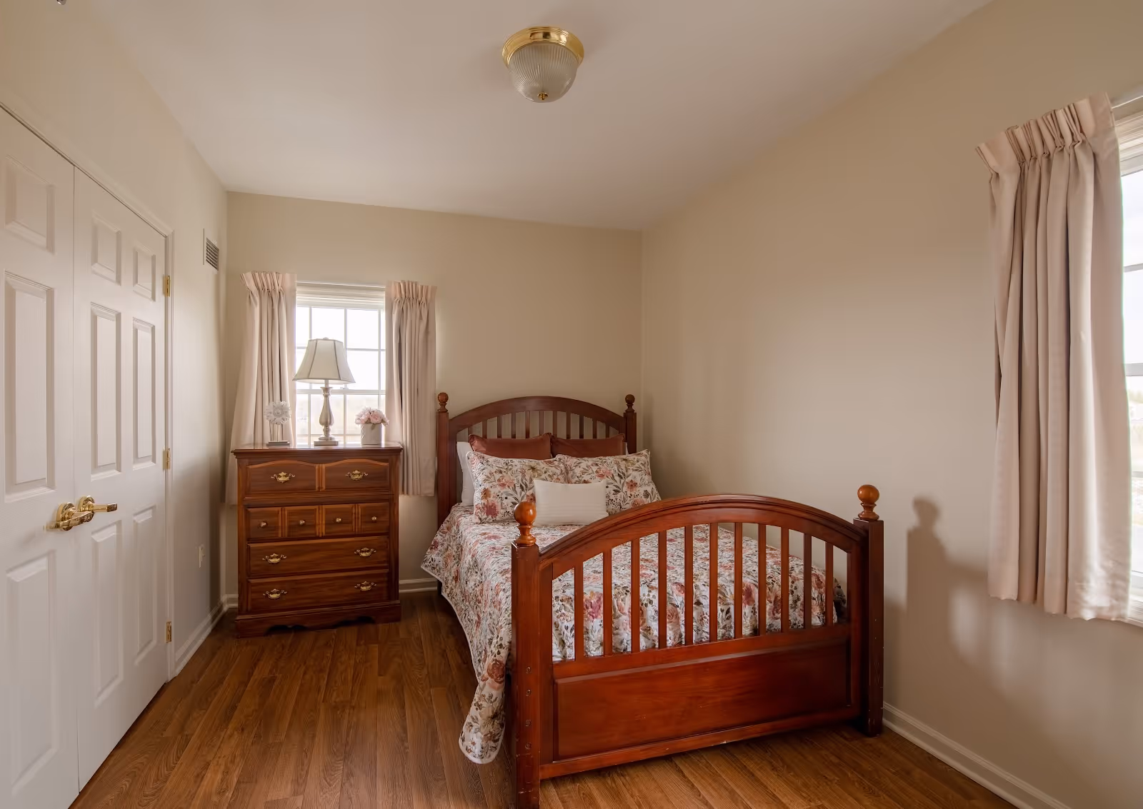 Cozy bedroom with a wooden single bed dressed in floral bedding, a matching wooden dresser with a lamp, hardwood floors, and curtained windows.