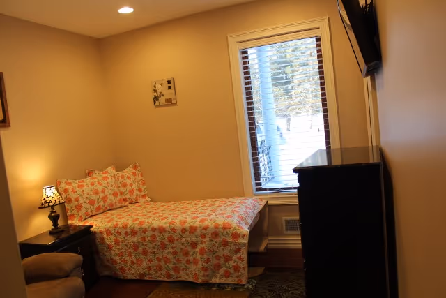 A small bedroom with a single bed covered in a floral bedspread and matching pillows. There is a nightstand with a decorative lamp next to the bed, a window with blinds letting in natural light, a dark dresser, and a wall-mounted TV.