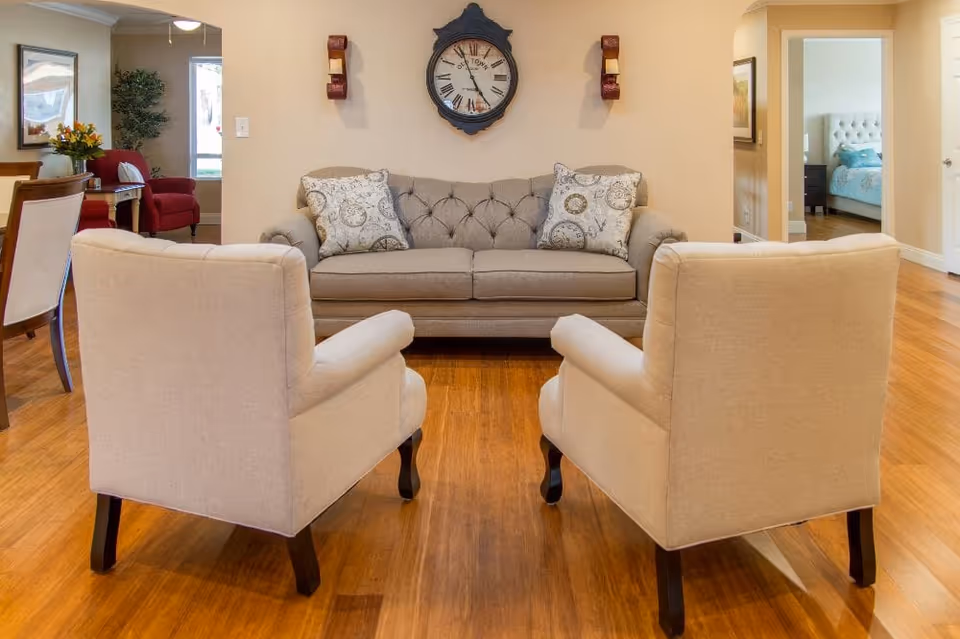 A cozy living room with a beige sofa adorned with two patterned pillows, two beige armchairs facing the sofa, a large wall clock above the sofa, wooden flooring, and a glimpse of a bedroom and dining area in the background.