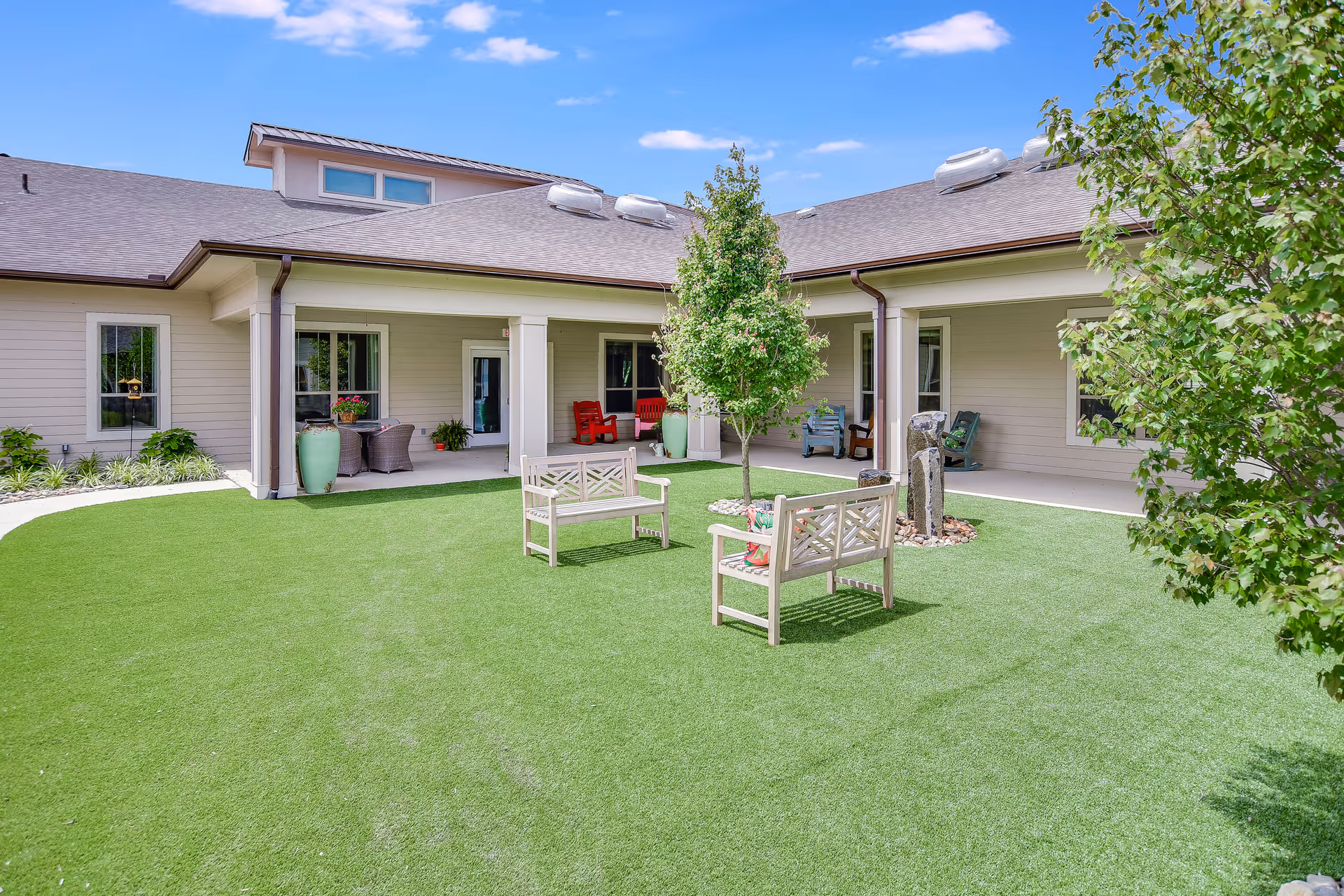 Outdoor courtyard area of a senior living facility with green artificial grass, two wooden benches, a small tree in the center, and a covered patio with chairs and potted plants. The building has beige siding and a brown roof under a blue sky with a few clouds.