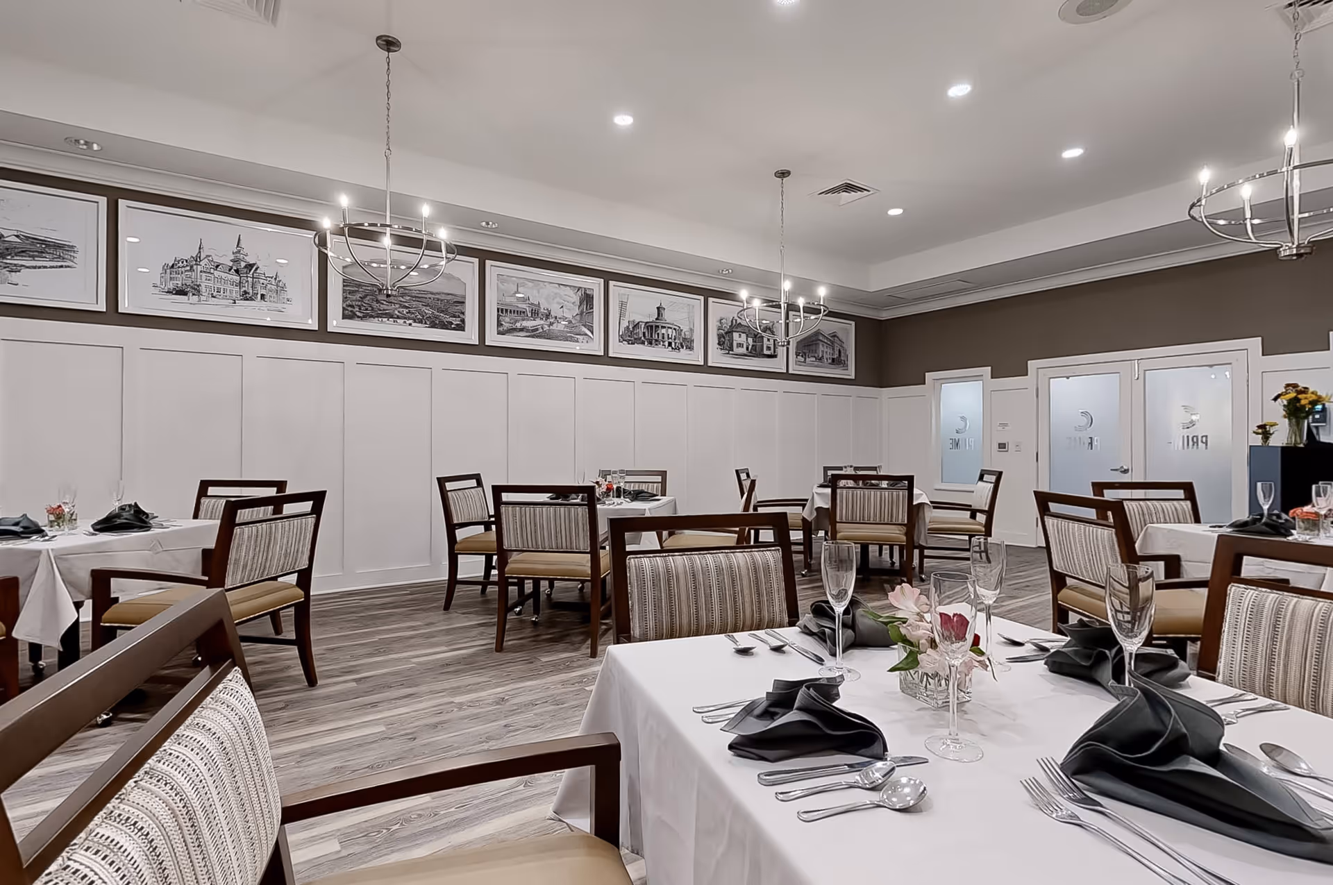 A dining room with several tables covered in white tablecloths, each set with silverware, black folded napkins, and glassware. The room has wooden chairs with striped cushions, wood flooring, and white paneled walls topped with framed black and white architectural sketches. Modern chandeliers hang from the ceiling, and frosted glass doors with the word 'PRIME' are visible in the background.