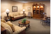 A cozy living room with two beige striped sofas, a round wooden coffee table with a plant on it, two table lamps on side tables, a wooden cabinet with shelves and decorative items, a framed picture on the wall, and a small round dining table with chairs.