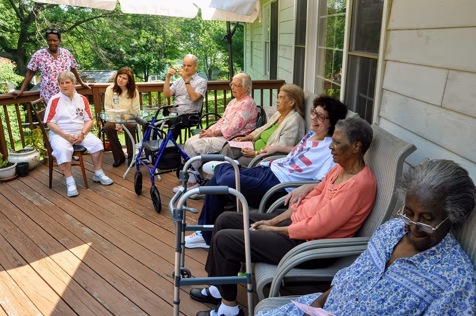A group of older adults seated on chairs on a wooden outdoor deck with walkers nearby and a staff member standing by, trees visible in the background.