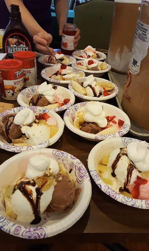 Several paper bowls of ice cream sundaes topped with whipped cream, fruit, and chocolate syrup on a table as a person adds toppings.