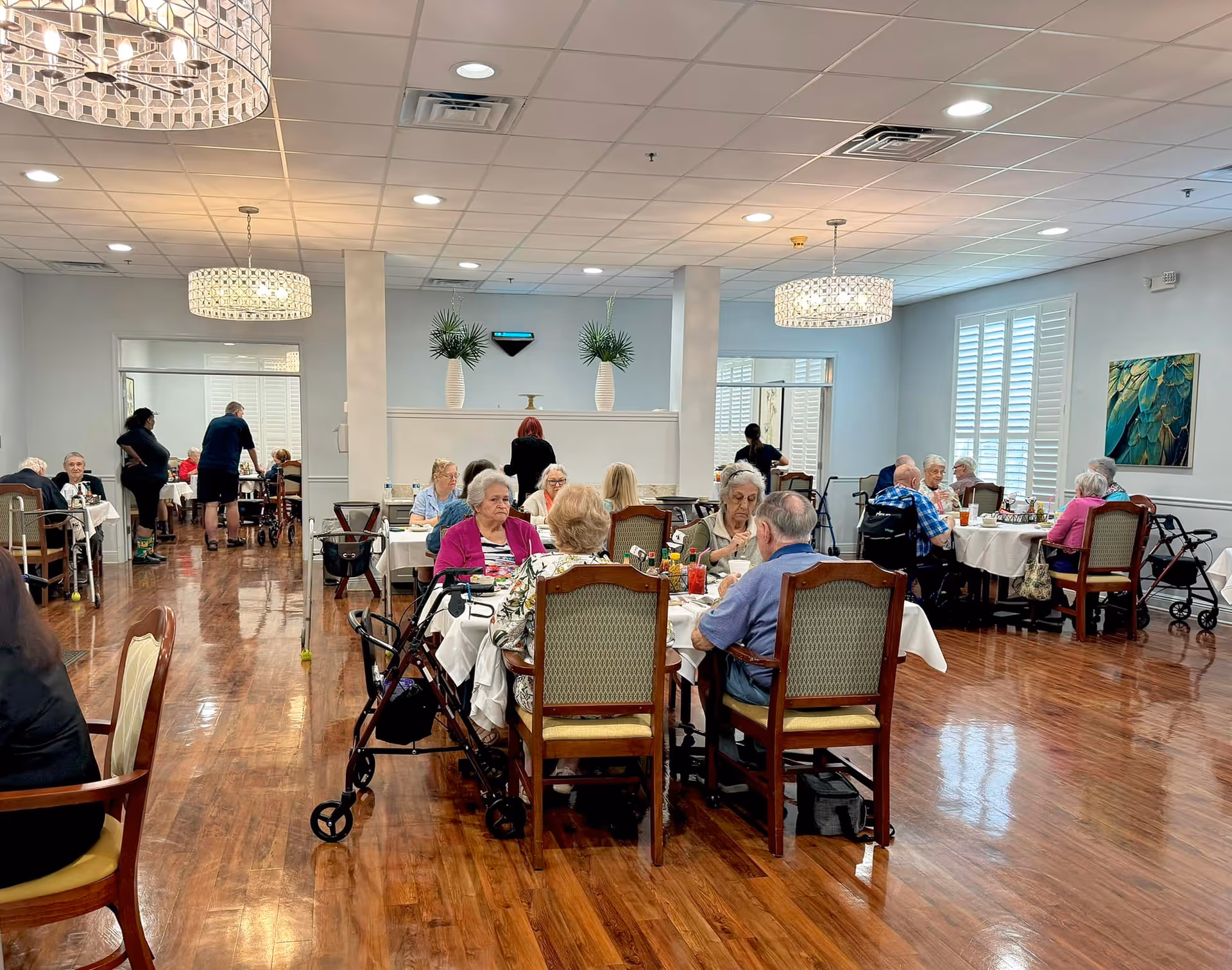 A dining room in The Windsor Senior Living Community with several elderly residents seated at tables enjoying a meal. The room has wooden floors, white tablecloths, and large windows with white shutters. There are chandeliers hanging from the ceiling and some decorative plants and artwork on the walls.