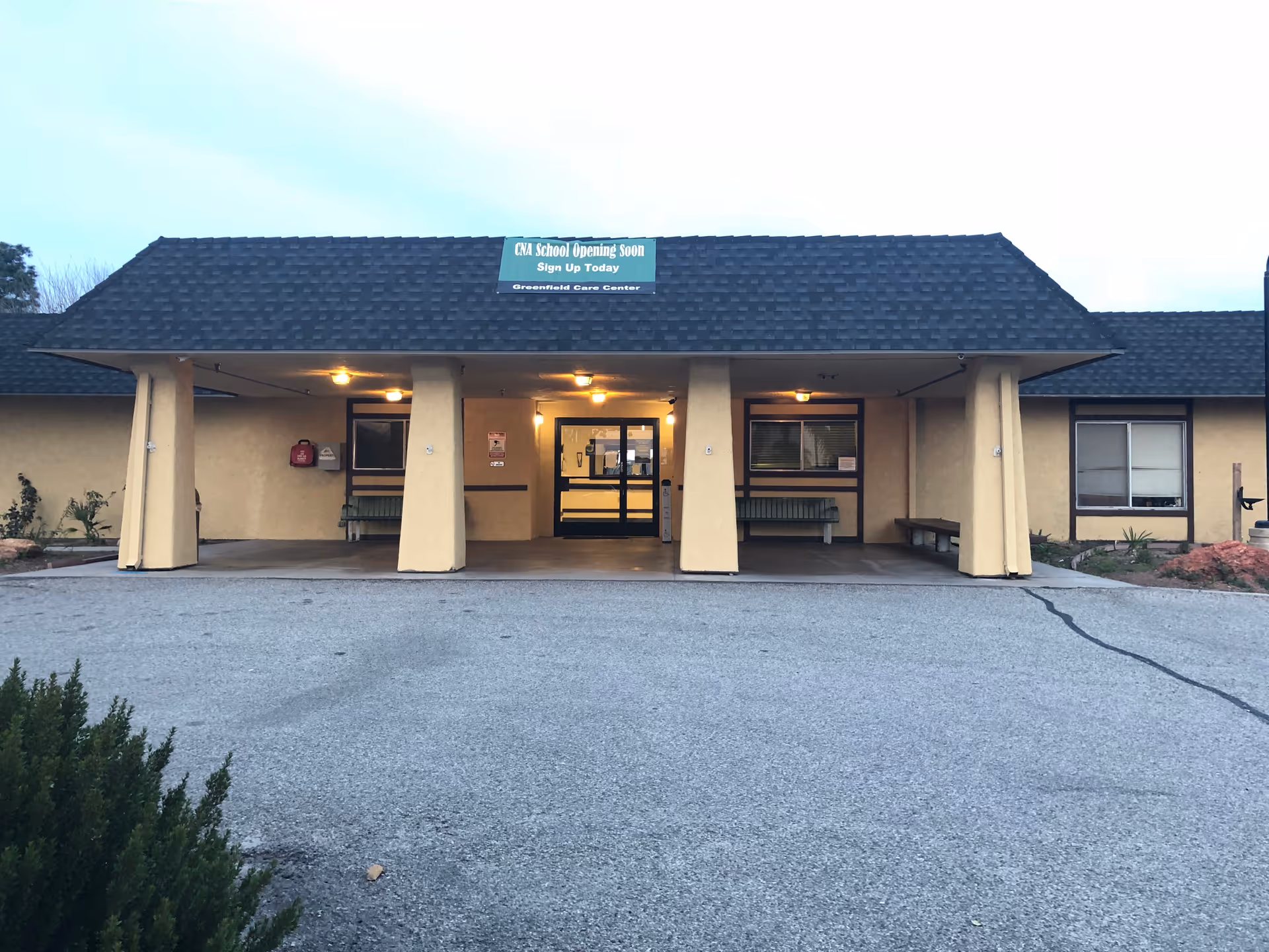 Front exterior entrance of a single-story care facility with a covered canopy, benches, and a rooftop sign.