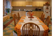 A dining room with a long table covered with a white tablecloth and an orange patterned runner. The table is set with placemats and a centerpiece featuring autumn-themed decorations. Wooden chairs surround the table. In the background, there is a kitchen area with light wood cabinets, a microwave, a refrigerator decorated with magnets, and a window with curtains.