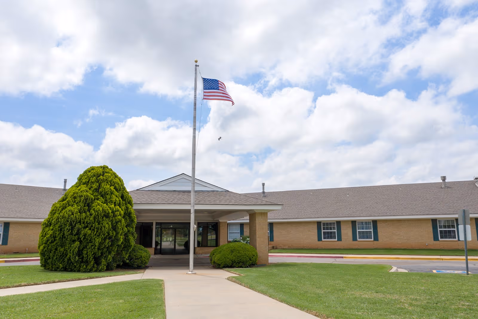 Front exterior view of Homestead Assisted Living of Clinton building with a covered entrance, an American flag on a flagpole, green bushes, and a well-maintained lawn under a partly cloudy sky.