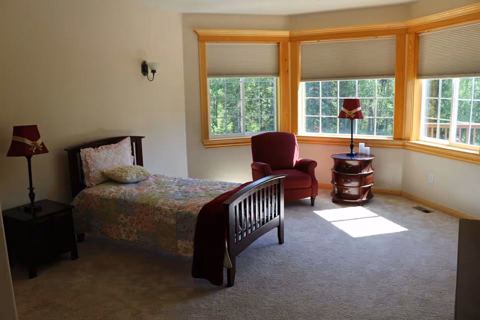 A bright bedroom with a single wooden bed covered with a floral quilt and pillows. There is a red armchair and a round wooden side table with a red lamp and some candles near three large windows with beige blinds. The room has beige carpet and light-colored walls with wooden trim around the windows.