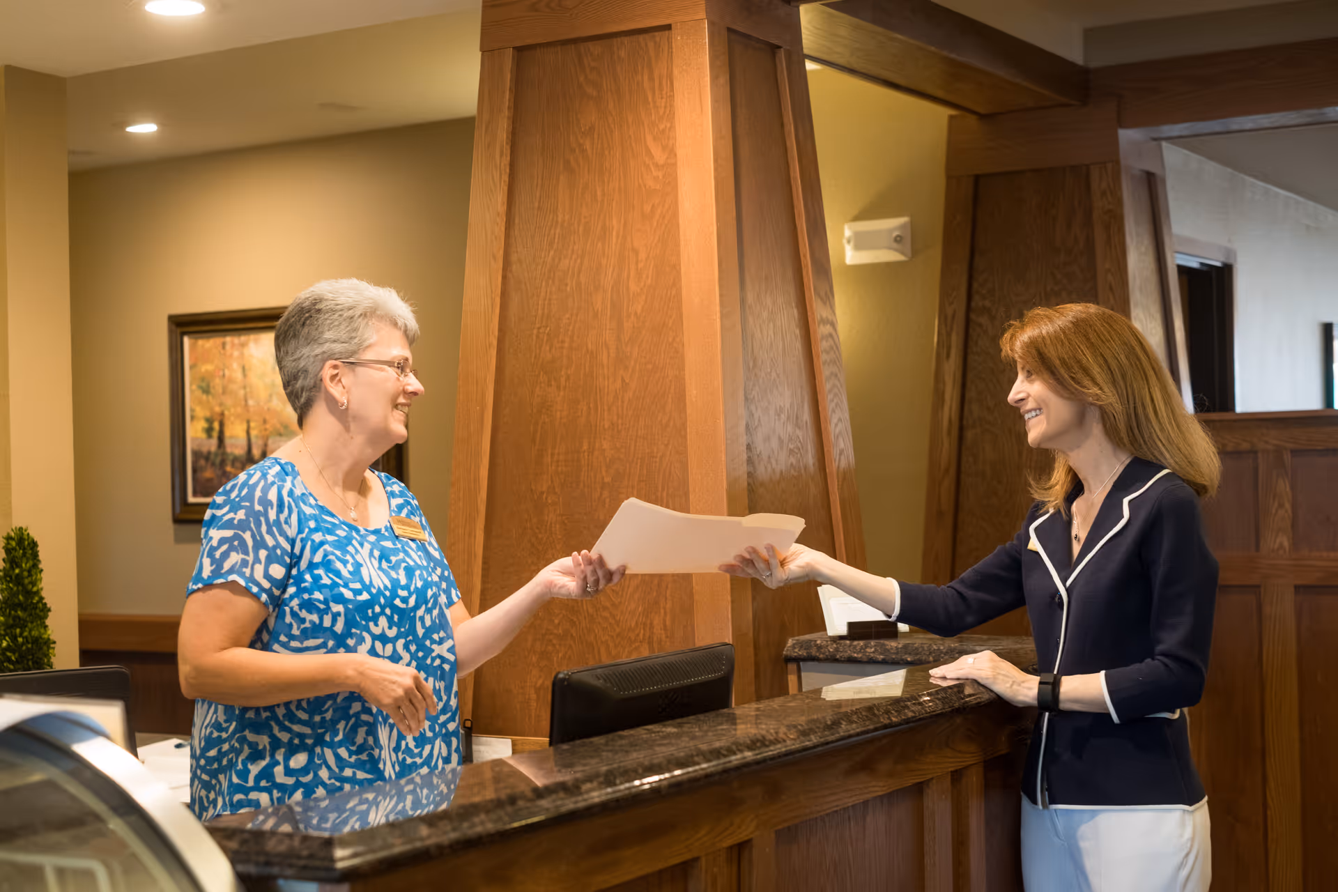 A woman in a blue patterned blouse stands behind a wooden reception desk handing a document to a smiling woman with long red hair wearing a navy blazer and white pants in a warmly lit interior space.