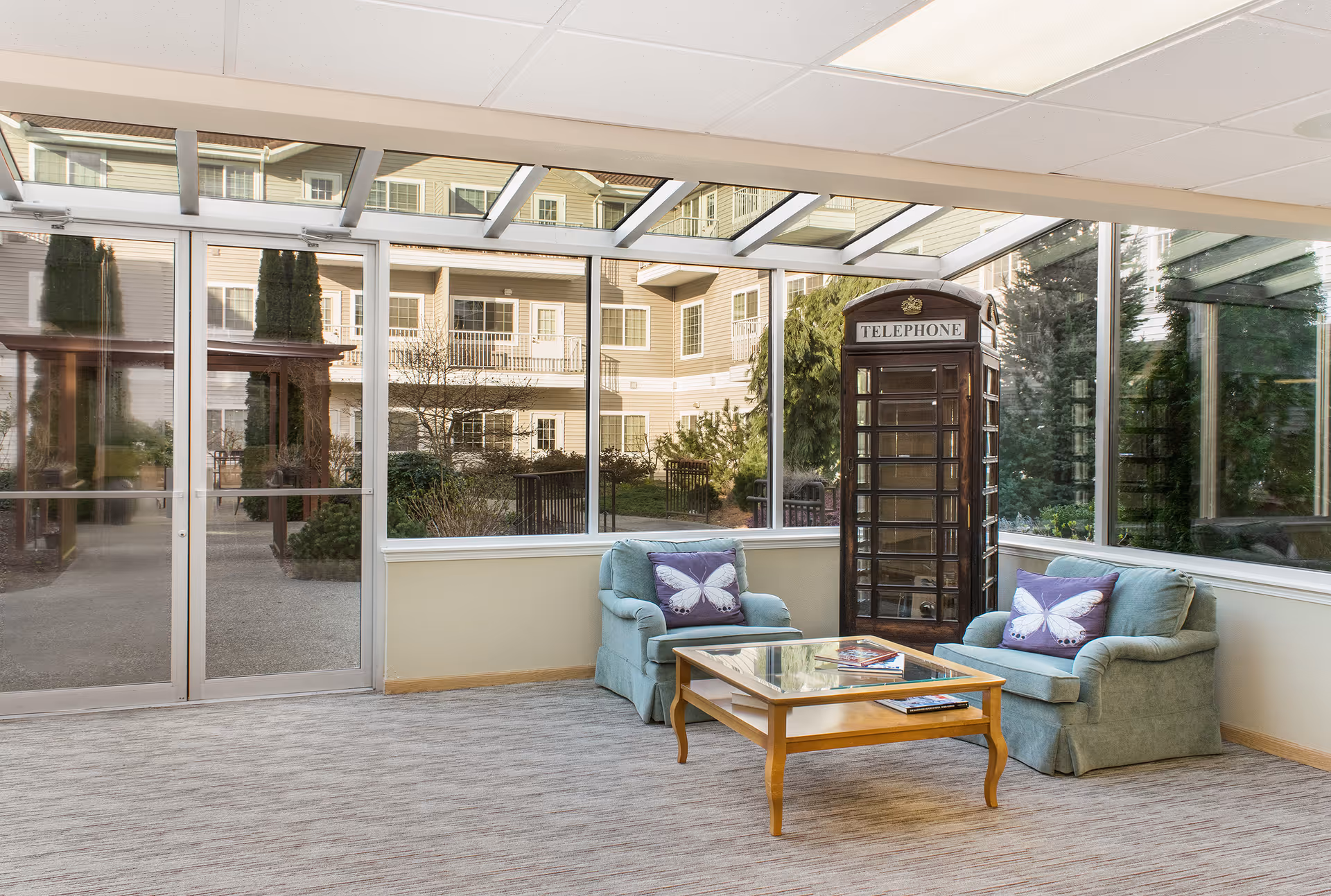 A bright indoor seating area with large windows showing an outdoor garden and building. The room contains two light blue armchairs with purple butterfly pillows, a wooden coffee table with magazines, and a vintage-style telephone booth in the corner.