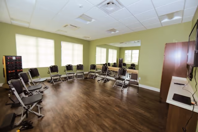 A spacious room with light green walls and wood flooring, arranged with folding chairs in a semi-circle facing a mirrored wall. There are windows with blinds letting in natural light, and a TV mounted on the wall with a cabinet below it. The room appears to be set up for group activities or meetings.