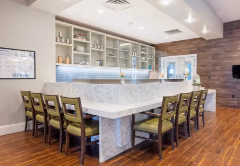 A modern communal dining area with a large marble countertop table surrounded by wooden chairs with green cushions. The background features open shelving with decorative items and a blue tiled backsplash. The floor is wooden, and the walls are light-colored with a wood accent wall on the right side.