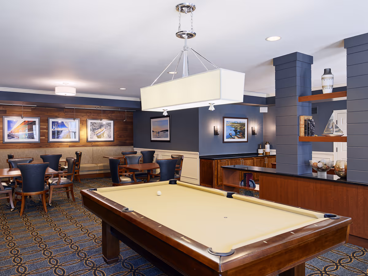 A recreational room featuring a pool table with a beige felt surface in the foreground. Behind the pool table, there are several wooden tables with black cushioned chairs arranged around them. The walls are painted dark blue with white wainscoting, and decorated with framed artwork. There is a wooden accent wall with additional framed pictures and a beige cushioned bench along it. The room has a patterned carpet and a large rectangular light fixture hanging above the pool table. Shelves with decorative items and cabinets are visible on the right side.
