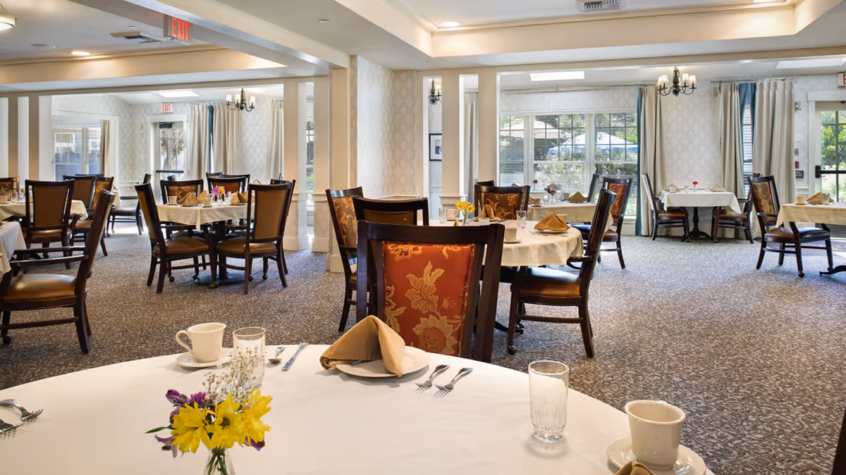 Bright, well-appointed dining room with round tables set for meals, chairs, and floral centerpieces.