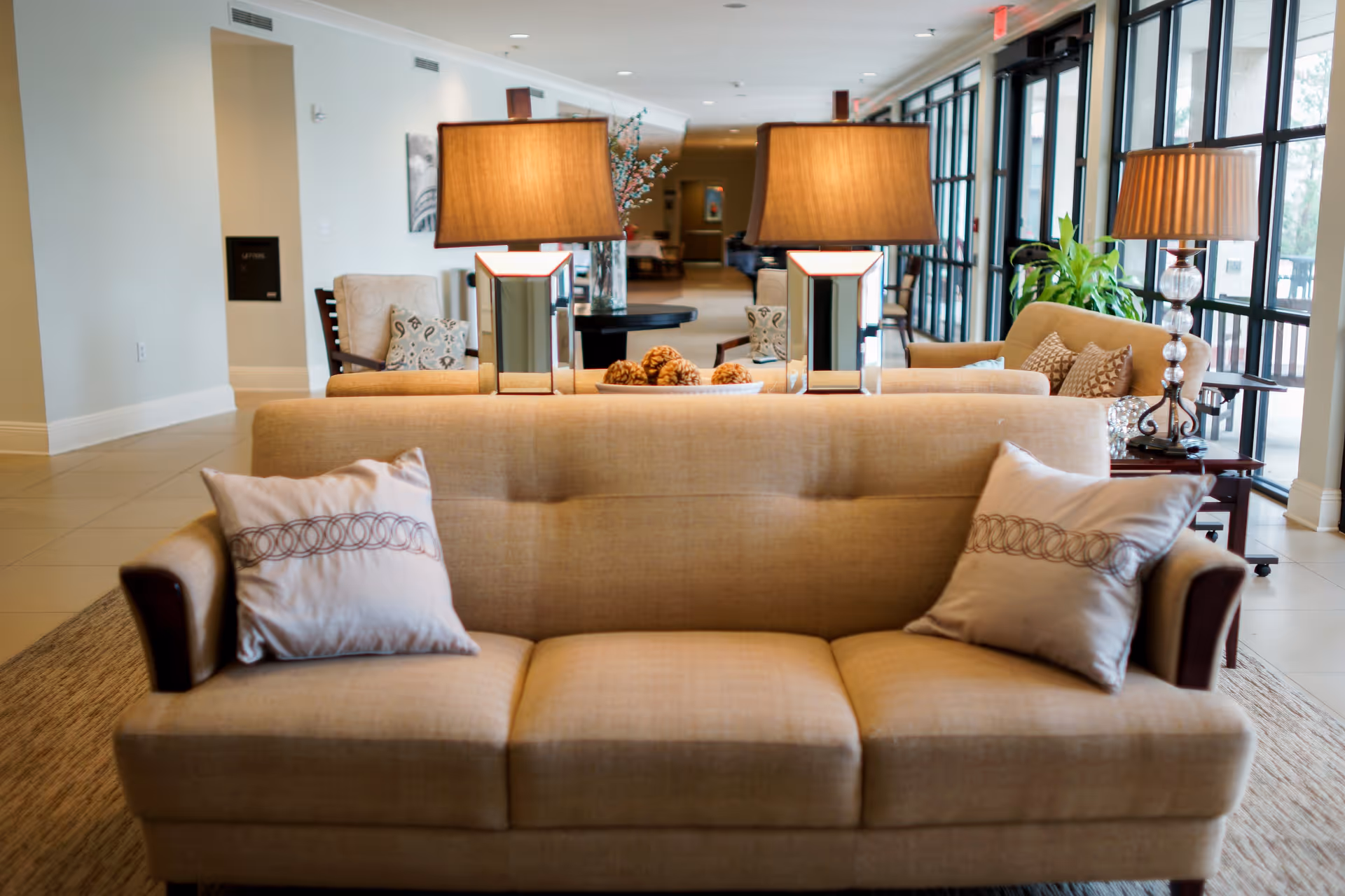 A bright communal lounge with beige sofas, decorative pillows, table lamps, and large windows along one side.