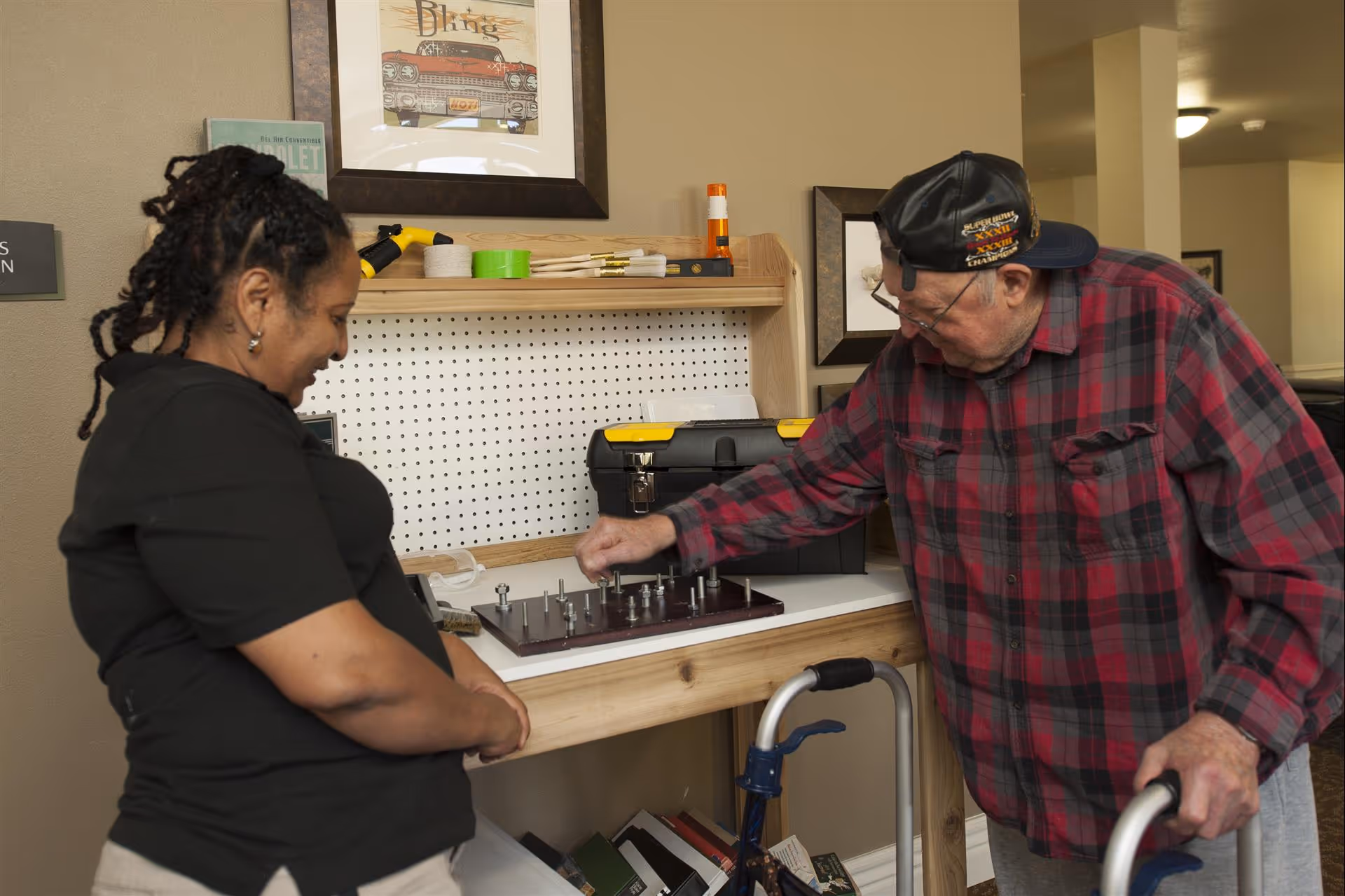 An elderly man using a walker is engaged in a tabletop activity involving bolts and screws, while a woman stands nearby watching and smiling. They are in a room with a wooden workbench, a toolbox, and framed pictures on the wall.