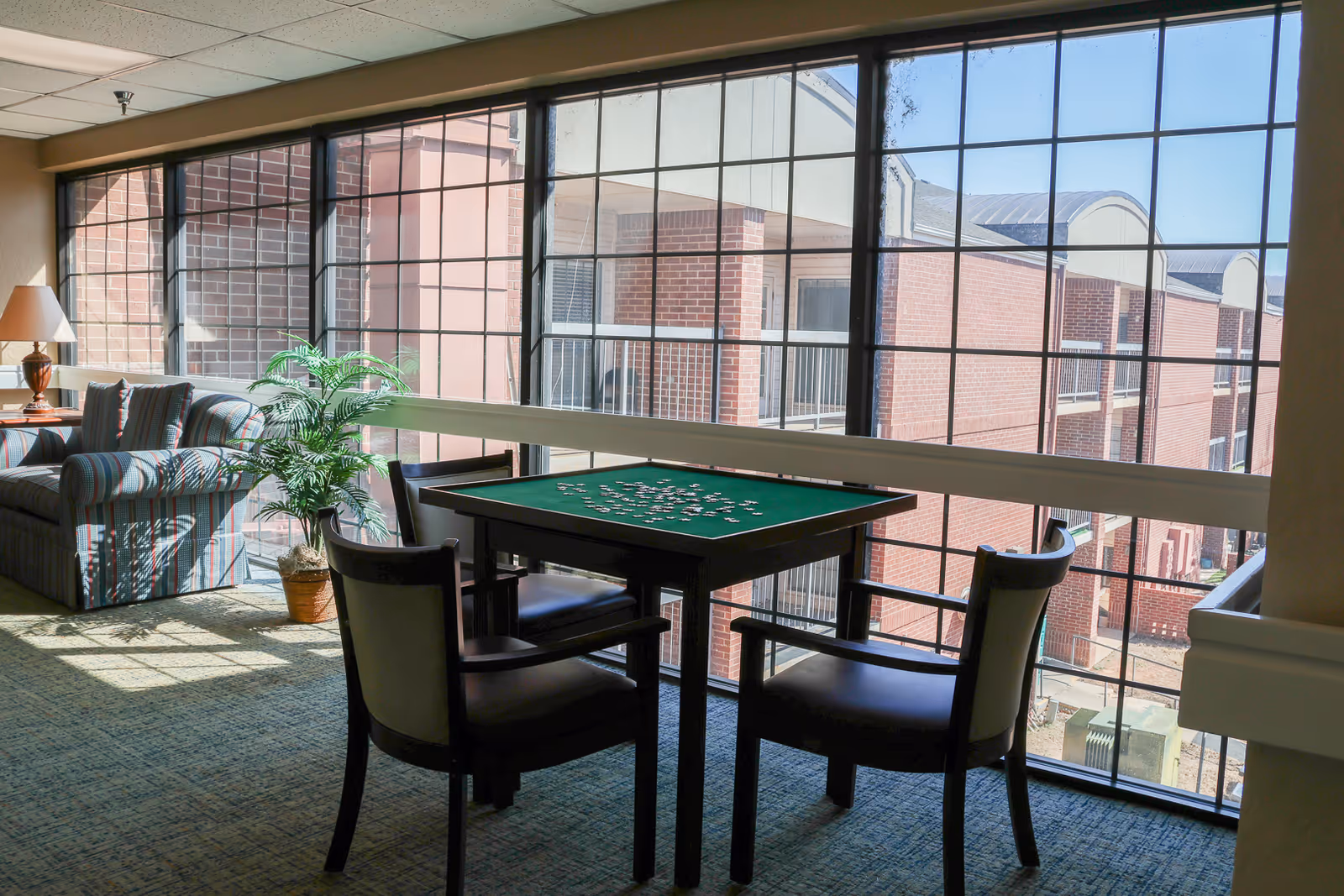 Sunlit senior living common area with a game table and chairs by a large grid window overlooking brick buildings, with a striped sofa and potted plant to the side.