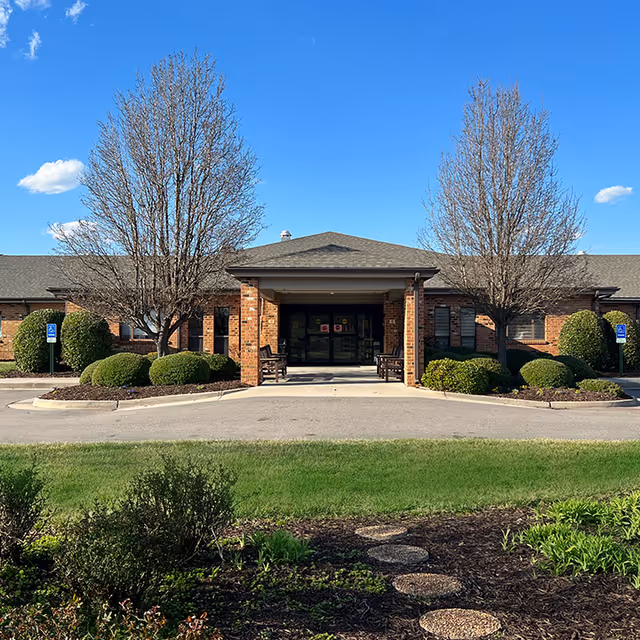 Front exterior view of a single-story brick building with a covered entrance, two leafless trees, trimmed bushes, and a clear blue sky.