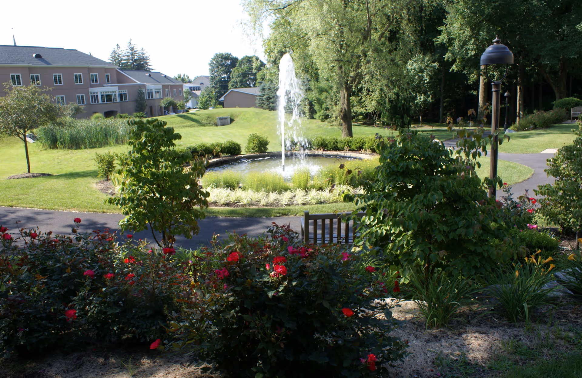 Landscaped outdoor courtyard with a central fountain, flowerbeds, walking paths, a bench, and a residential building in the background.