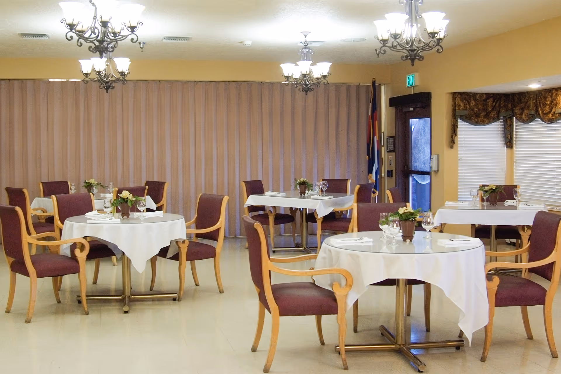 A dining room with round and rectangular tables covered with white tablecloths, each set with glasses, napkins, and small flower arrangements. The room has wooden chairs with purple cushions, chandeliers hanging from the ceiling, and large windows with blinds and decorative valances.