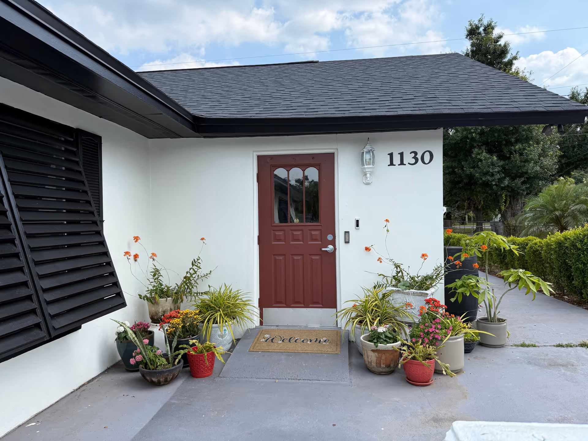 Front entrance of a building with a maroon door, white walls, and the number 1130 displayed beside the door. There are multiple potted plants with flowers arranged on both sides of the door, and a welcome mat is placed in front of the door. The building has a black roof and black window shutters.