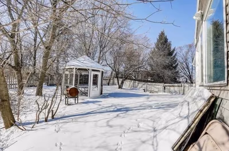 Snow-covered backyard with a glass gazebo, trees, and a fenced area beside a building